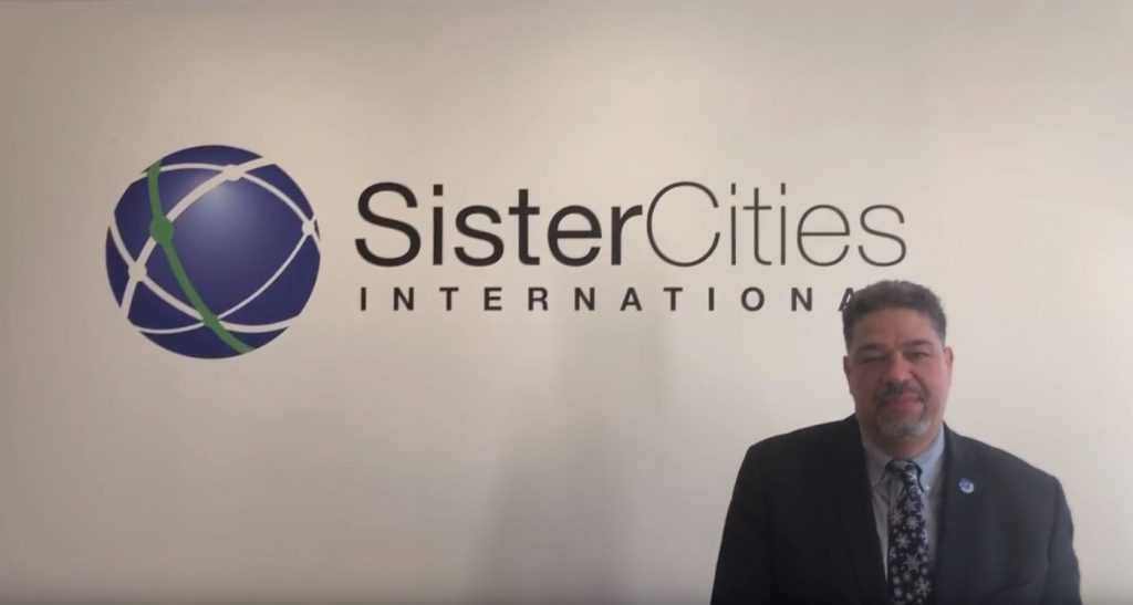 Roger-Mark De Souza stands in front of the Sister Cities International logo